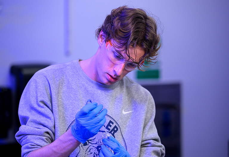 Science student in lab in grey sweatshirt and safety goggles, blue gloves